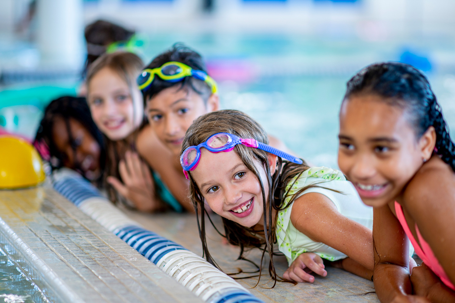 A multi-ethnic group of kids are indoors in a pool. Some of them are wearing goggles and smiling.