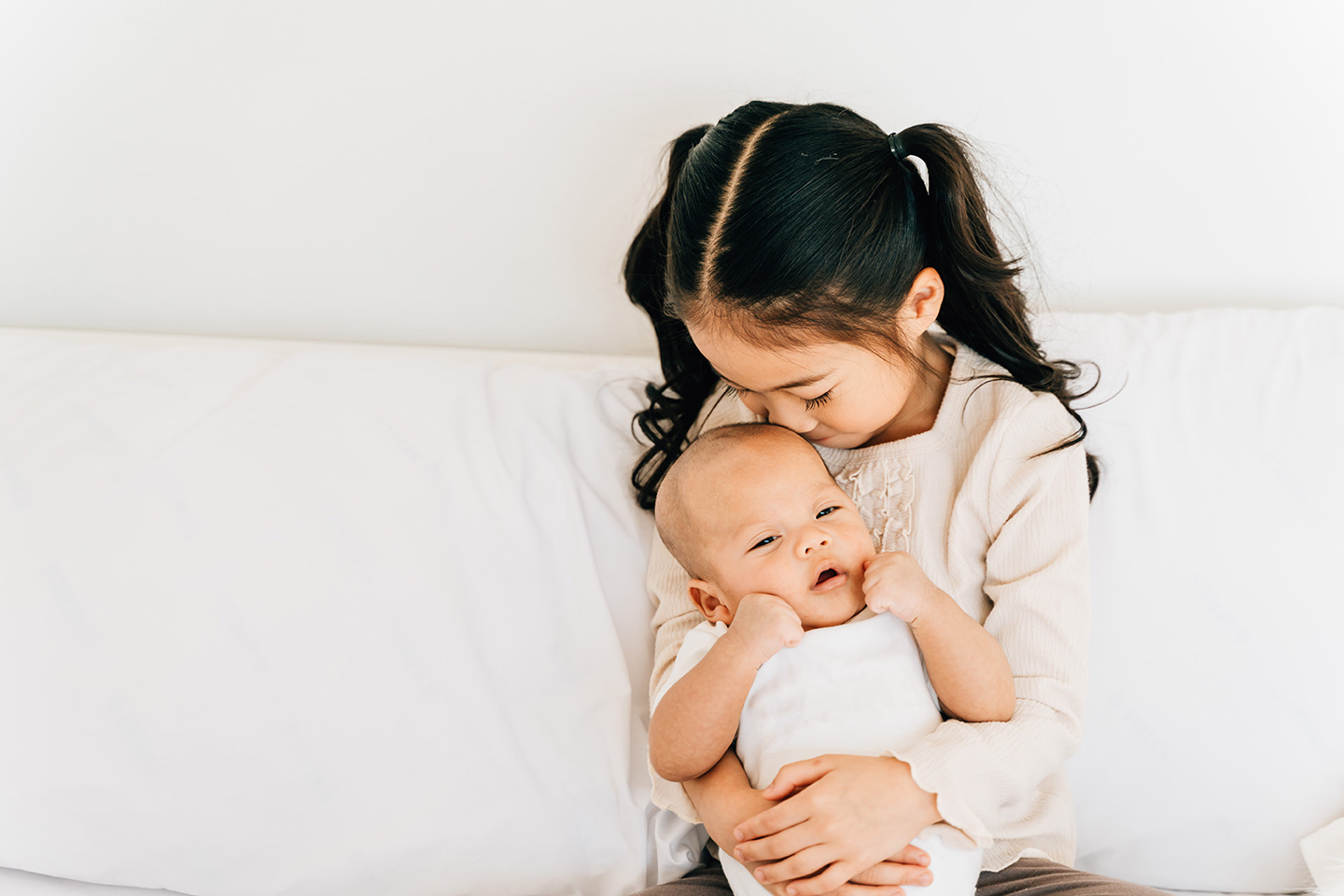 Caring Asian girl in casual wear hugging small brother embracing newborn baby with opened mouth smelling toddler sitting on bed in house