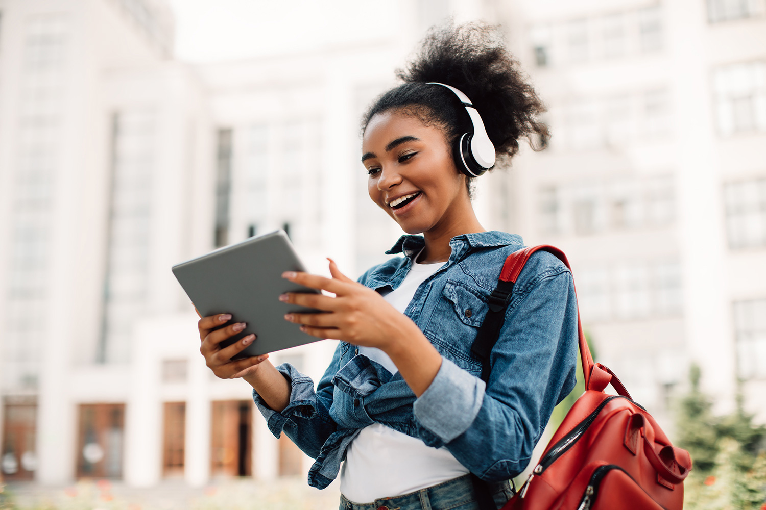 Excited African American Student Girl Using Digital Tablet Wearing Headphones Standing Near University Building Outdoors.