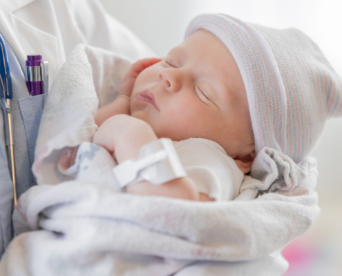 A Caucasian female infant is asleep in the arms of a pediatrician. The baby is swaddled in a blanket and is wearing a warm cap.